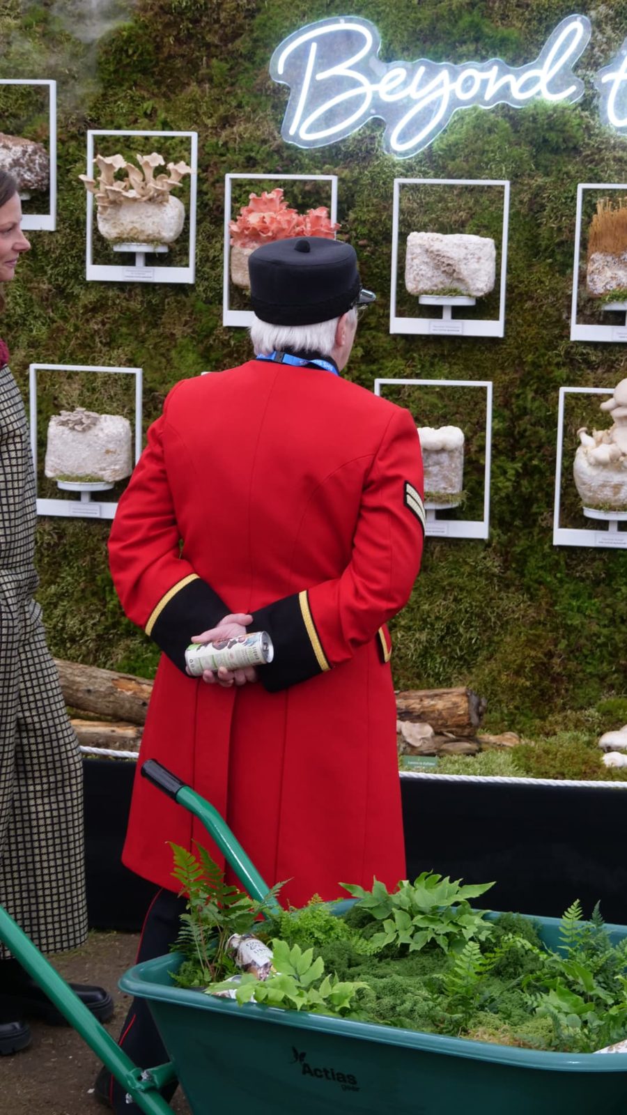 A Chelsea Pensioner in a red coat holding a Grass and Co. can in front of the Beyond the Button mushroom wall